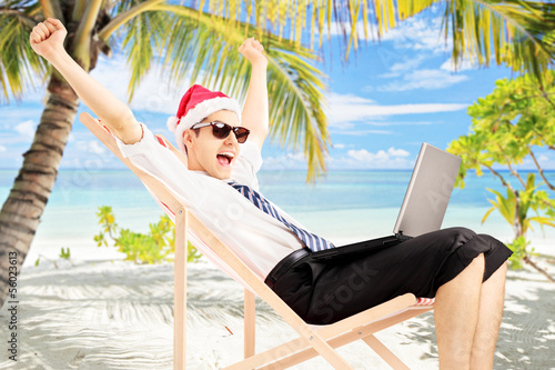 Photo: Excited male with santa hat working on a laptop, on a beach Excited male with santa hat working on a laptop, on a beach