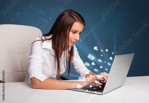 Photo: Young lady sitting at dest and typing on laptop with message ico Young lady sitting at dest and typing on laptop with message ico