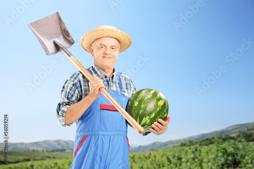 Photo: Smiling farmer holding a watermelon and shovel at field Smiling farmer holding a watermelon and shovel at field