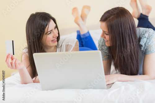 Photo: Pretty friends using a laptop to shop online lying on bed Pretty friends using a laptop to shop online lying on bed