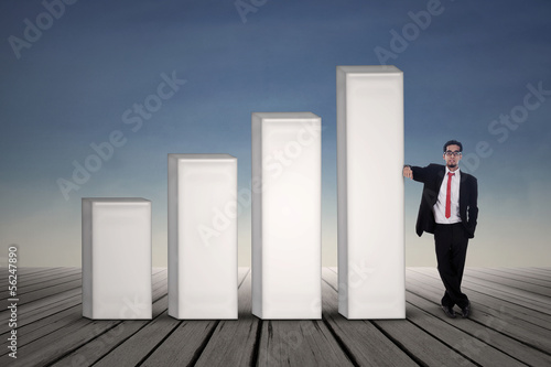 Photo: Asian businessman in black suit standing next to bar chart Asian businessman in black suit standing next to bar chart