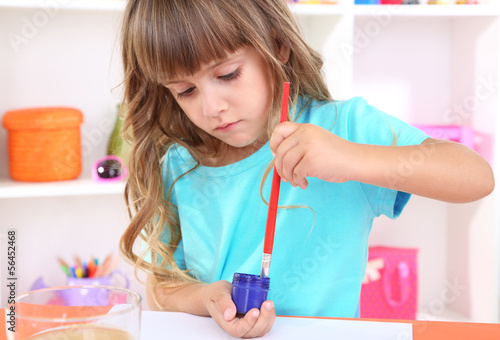 Photo: Little girl draws sitting at table in room on shelves Little girl draws sitting at table in room on shelves