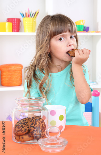 Photo: Little girl eating cookies sitting at table in room Little girl eating cookies sitting at table in room