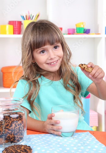 Photo: Little girl eating cookies and drinking milk sitting at table Little girl eating cookies and drinking milk sitting at table