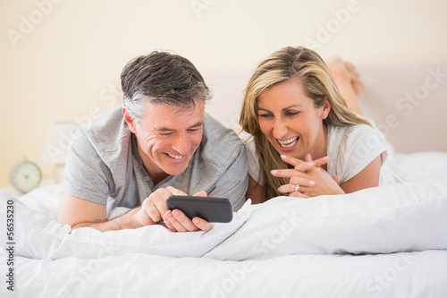 Photo: Happy couple lying on a bed and watching a mobile phone Happy couple lying on a bed and watching a mobile phone