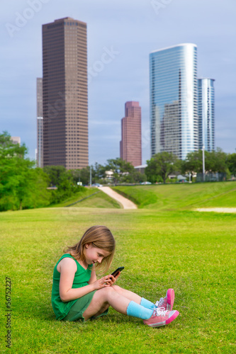 Photo: Blond kid girl playing with smartphone sitting on park lawn at c Blond kid girl playing with smartphone sitting on park lawn at c