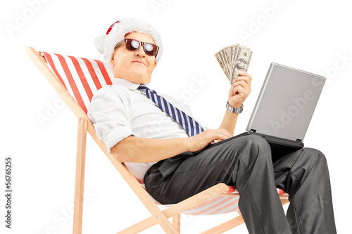 Photo: Senior man with santa hat on a chair holding banknotes and pc Senior man with santa hat on a chair holding banknotes and pc