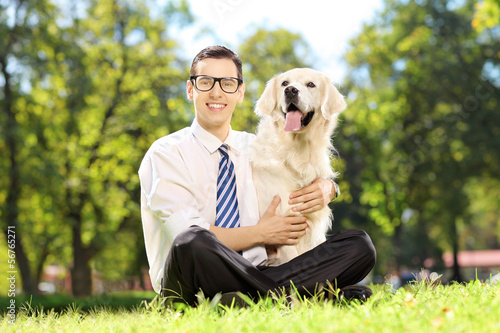 Photo: Smiling male with glasses sitting on a grass and hugging his dog Smiling male with glasses sitting on a grass and hugging his dog
