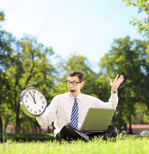 Photo: Angry man with laptop sitting on grass and looking at clock Angry man with laptop sitting on grass and looking at clock