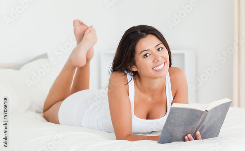 Photo: cute woman lying on bed with book looking into camera cute woman lying on bed with book looking into camera