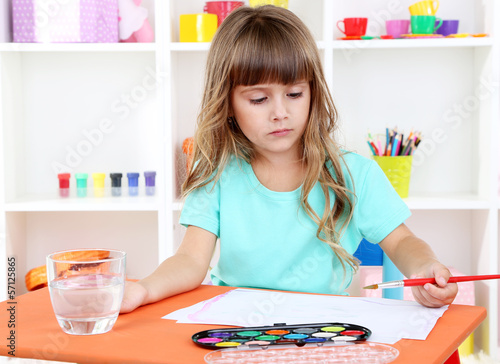 Photo: Little girl draws sitting at table in room on shelves Little girl draws sitting at table in room on shelves