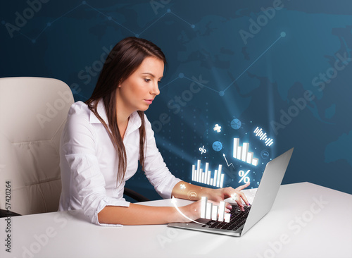 Photo: Pretty woman sitting at desk and typing on laptop with diagrams Pretty woman sitting at desk and typing on laptop with diagrams