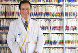 Photo: Portrait of smiling doctor in lab coat standing in front of filed medical records on shelf Portrait of smiling doctor in lab coat standing in front of filed medical records on shelf