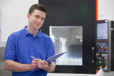 Photo: Portrait of confident technician with clipboard in front of lathe cutting machine in hi-tech manufacturing plant Portrait of confident technician with clipboard in front of lathe cutting machine in hi-tech manufacturing plant