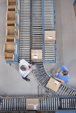 Photo: Businessman with digital tablet and worker packing box at conveyor belt in distribution warehouse Businessman with digital tablet and worker packing box at conveyor belt in distribution warehouse