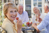 Photo: Portrait of smiling senior woman eating strawberry on patio with friends enjoying wine and lunch in background Portrait of smiling senior woman eating strawberry on patio with friends enjoying wine and lunch in background