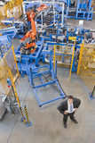 Photo: Portrait of confident businessman in front of robotic machinery lifting steel fencing on production line in manufacturing plant Portrait of confident businessman in front of robotic machinery lifting steel fencing on production line in manufacturing plant