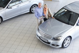 Photo: Portrait of smiling couple with brochure looking at car in car dealership showroom Portrait of smiling couple with brochure looking at car in car dealership showroom