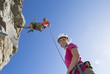 Photo: Portrait of woman looking at camera as male rock climber is abseiling down rock face Portrait of woman looking at camera as male rock climber is abseiling down rock face