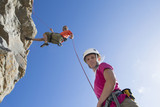 Photo: Portrait of woman looking at camera as male rock climber is abseiling down rock face Portrait of woman looking at camera as male rock climber is abseiling down rock face
