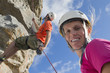 Photo: Portrait of smiling woman looking at camera as male rock climber is abseiling down rock face Portrait of smiling woman looking at camera as male rock climber is abseiling down rock face