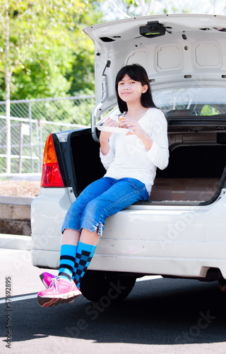Photo: Preteen girl sitting on back car bumper eating lunch Preteen girl sitting on back car bumper eating lunch
