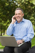Photo : Laughing professor talking on phone while sitting on bench using laptop on campus Laughing professor talking on phone while sitting on bench using laptop on campus