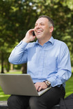 Photo : Laughing professor talking on phone while sitting on bench using laptop on campus Laughing professor talking on phone while sitting on bench using laptop on campus