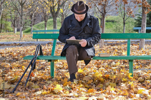 Photo: Elderly man on crutches using a tablet in the park Elderly man on crutches using a tablet in the park