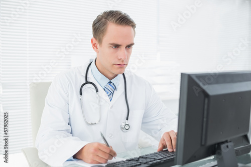 Photo: Doctor writing a note while using computer at medical office Doctor writing a note while using computer at medical office
