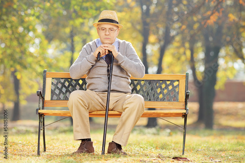 Photo: Thoughtful senior man with a cane sitting on bench in a park Thoughtful senior man with a cane sitting on bench in a park