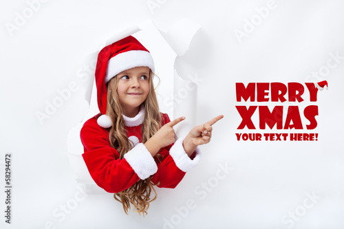 Photo: Santa girl pointig to copy space - leaning through hole in paper Santa girl pointig to copy space - leaning through hole in paper
