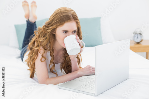 Photo: Casual blond drinking coffee while using laptop in bed Casual blond drinking coffee while using laptop in bed
