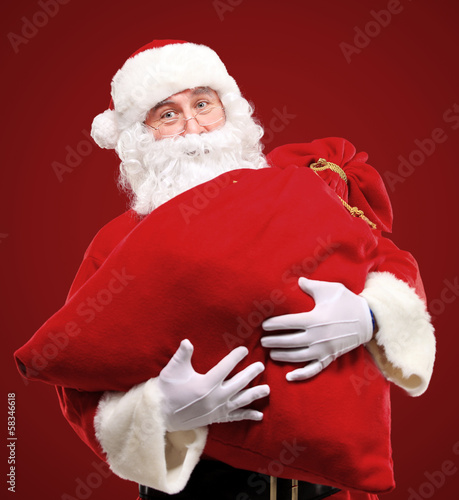 Photo: Portrait of Santa Claus embracing huge red sack with gifts Portrait of Santa Claus embracing huge red sack with gifts