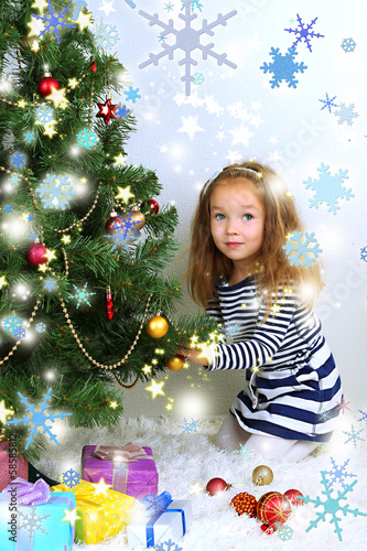 Photo: Little girl decorating Christmas tree with baubles in room Little girl decorating Christmas tree with baubles in room
