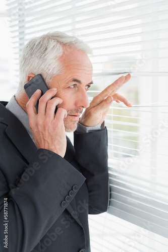 Photo: Businessman peeking through blinds while on call in office Businessman peeking through blinds while on call in office