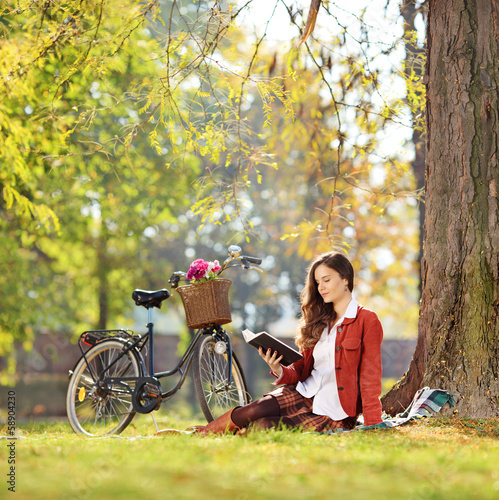 Photo: Young beautiful female with her bike, reading a novel in park Young beautiful female with her bike, reading a novel in park