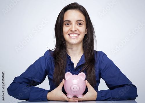 Photo: Happy smiling woman holding a piggy bank against gray background Happy smiling woman holding a piggy bank against gray background