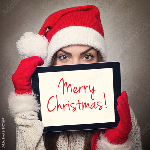 Photo: Cute young woman hiding behind tablet computer wearing Santa hat Cute young woman hiding behind tablet computer wearing Santa hat