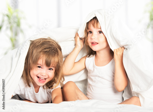 Photo: happy little girls twins sister in bed under the blanket having happy little girls twins sister in bed under the blanket having