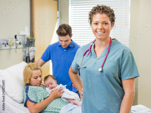 Photo: Nurse Standing With Couple And Newborn Baby In Background Nurse Standing With Couple And Newborn Baby In Background