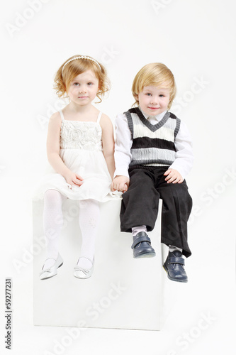 Photo: Little happy girl and boy sit on white big cube on white Little happy girl and boy sit on white big cube on white