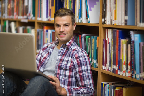 Photo: Smiling young student sitting on library floor using laptop Smiling young student sitting on library floor using laptop