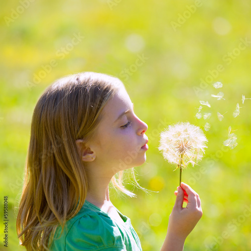 Photo: Blond kid girl blowing dandelion flower in green meadow Blond kid girl blowing dandelion flower in green meadow