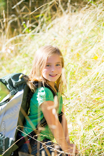 Photo: Blond explorer kid girl walking with backpack in grass Blond explorer kid girl walking with backpack in grass