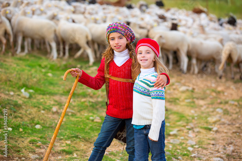 Photo: Kid girl shepherdess sisters happy with flock of sheep and stick Kid girl shepherdess sisters happy with flock of sheep and stick
