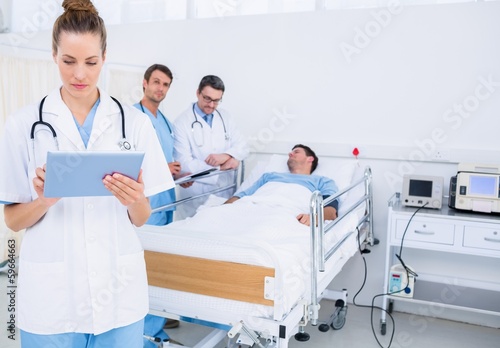 Photo: Doctor using digital tablet with colleagues and patient behind Doctor using digital tablet with colleagues and patient behind