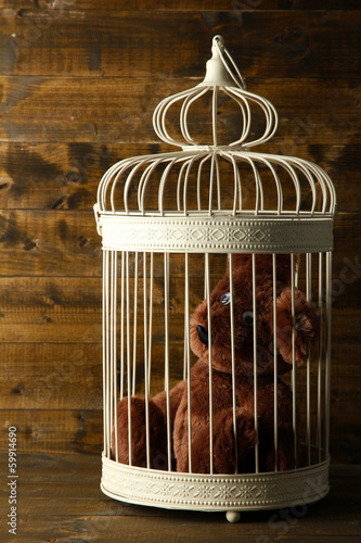 Photo: Toy bear in decorative cage on wooden background Toy bear in decorative cage on wooden background