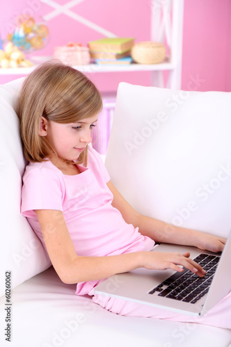 Photo: Little girl sitting with laptop on sofa in room Little girl sitting with laptop on sofa in room