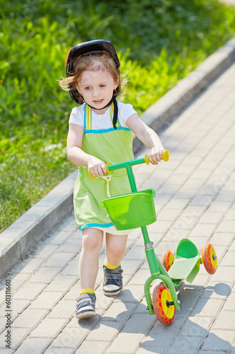 Photo: Little girl with three-wheeled scooter goes along alley in park Little girl with three-wheeled scooter goes along alley in park